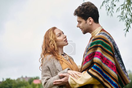 Un hombre y una mujer, vestidos al estilo boho, parados uno al lado del otro en un exuberante parque verde, en una cita romántica.