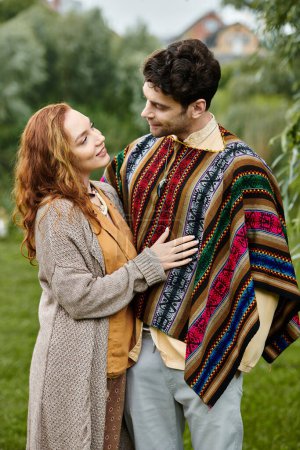 A man in boho style clothing stands beside a woman wrapped in a blanket in a serene green park setting.