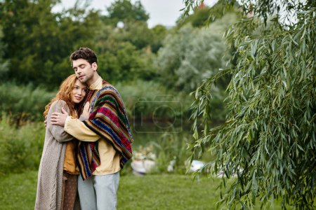 A man and a woman in boho style clothes stand closely together in a green park, exuding an air of romance and elegance.