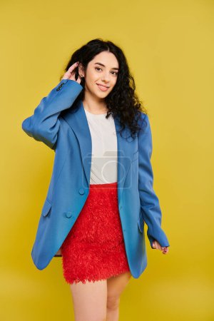 Foto de Mujer morena joven con el pelo rizado posa en elegante chaqueta azul y falda roja, exudando confianza y estilo sobre fondo amarillo. - Imagen libre de derechos