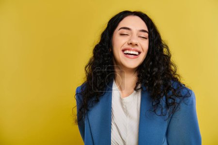 Téléchargez les photos : Une jeune femme aux cheveux bouclés sourit et porte une veste bleue, rayonnant de joie et de confiance dans un décor de studio avec une toile de fond jaune. - en image libre de droit