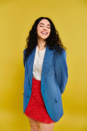 Photo for Young brunette woman with curly hair poses in stylish blue jacket and red skirt against yellow backdrop, showcasing her emotions. - Royalty Free Image
