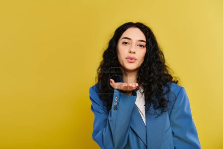 A young brunette woman with curly hair elegantly posing in a blue jacket, making a hand gesture, against a vibrant yellow backdrop.