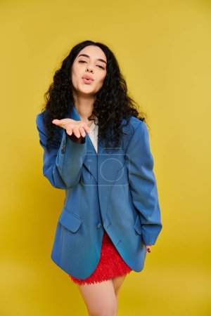 Téléchargez les photos : Jeune femme brune aux cheveux bouclés frappe une pose dans une veste bleue élégante et une jupe rouge, exsudant une énergie vibrante sur fond jaune. - en image libre de droit