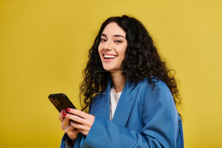 Téléchargez les photos : Une jeune femme brune aux cheveux bouclés dans une robe bleue tenant avec confiance un téléphone portable, respirant style et sophistication. - en image libre de droit