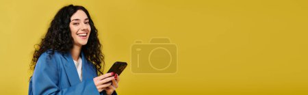 Photo for An expressive young brunette woman with curly hair smiling while holding a smartphone, exuding joy in a studio setting with a yellow background. - Royalty Free Image