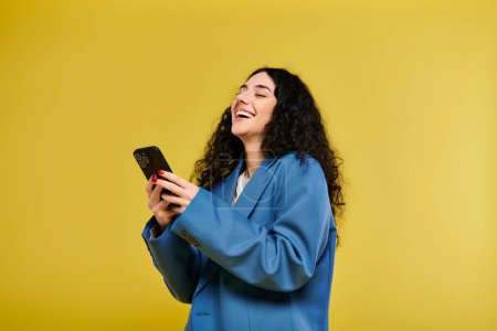 Téléchargez les photos : Une jeune femme élégante avec des cheveux bouclés dans une veste bleue tient un téléphone portable sur un fond jaune vif. - en image libre de droit