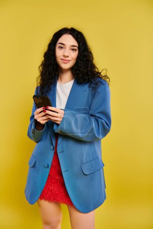 Stylish young woman with curly hair and trendy outfit taking a selfie with her cell phone in front of a vibrant yellow wall.