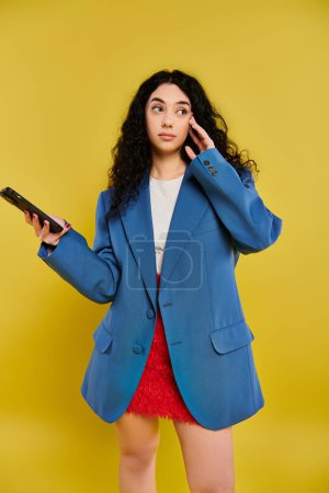 A young brunette woman with curly hair, dressed in a blue jacket, holding a cell phone in a vibrant yellow studio.