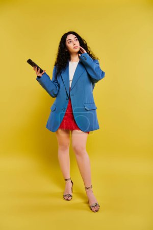 Téléchargez les photos : Jeune femme brune aux cheveux bouclés dans un blazer bleu, tenant avec confiance un téléphone portable sur fond jaune vif. - en image libre de droit