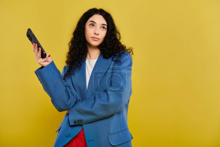 Brunette woman with curly hair in a blue jacket stands, holding a smartphone, exhibiting elegance and control.