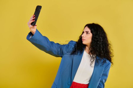 Téléchargez les photos : Une jeune femme élégante aux cheveux bouclés capture un moment franc avec son téléphone portable sur fond jaune ensoleillé. - en image libre de droit