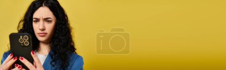 Foto de Mujer morena con el pelo largo y rizado mirando atentamente a su teléfono celular, mostrando una mezcla de emociones, contra un vibrante telón de fondo amarillo. - Imagen libre de derechos