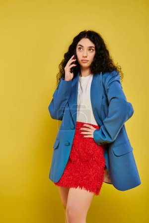 Young brunette woman with curly hair posing stylishly in a blue jacket and red skirt against a yellow studio background.