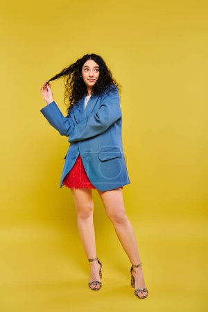 Photo for A brunette woman with curly hair posing in a blue jacket and red skirt, exuding style and charm against a vibrant yellow backdrop. - Royalty Free Image
