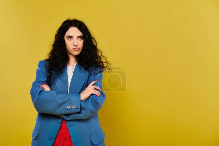 Photo for A young brunette woman with curly hair confidently stands with her arms crossed in front of a vibrant yellow background. - Royalty Free Image