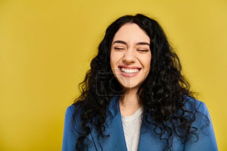 A brunette woman with curly hair joyfully smiling while wearing a blue jacket against a yellow background.
