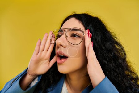 A young, brunette woman with curly hair and glasses making a silly expression in a stylish outfit against a yellow backdrop.