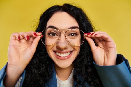 Téléchargez les photos : Jeune femme brune aux cheveux bouclés, portant des lunettes, sourit vivement à la caméra dans une tenue élégante sur fond jaune. - en image libre de droit