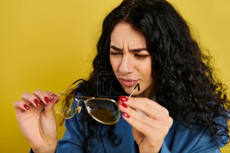 Une jeune femme élégante aux cheveux bouclés tient une paire de lunettes de soleil, mettant en valeur son style ludique et sans effort sur fond jaune.