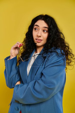 Photo for A young brunette woman with curly hair poses in a stylish blue jacket, showing glasses in a studio with a yellow background. - Royalty Free Image