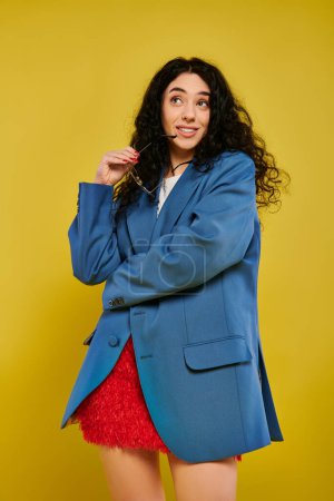 Photo for A young brunette woman with curly hair poses in a stylish blue jacket and red skirt, expressing emotions against a yellow background. - Royalty Free Image