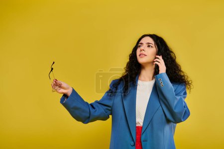 A stylish young woman with curly hair wears a blue jacket and talks on a cell phone in a vibrant studio setting.
