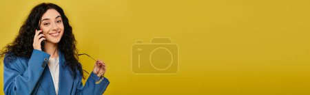 Photo for A young brunette woman with curly hair wearing a blue jacket, talking on a cell phone in a studio with a yellow background. - Royalty Free Image