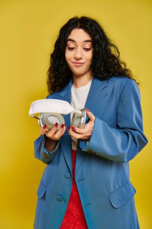 A young brunette woman with curly hair poses in a studio setting, showcasing her emotions as she holds a white headphones in her hands against a yellow background.
