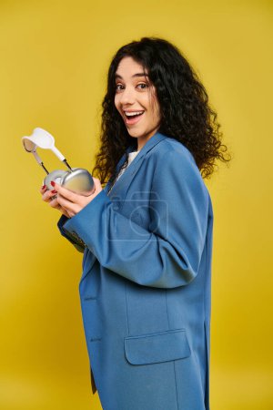 A young brunette woman in a blue jacket holds headphones, expressing curiosity, in a studio with a yellow background.