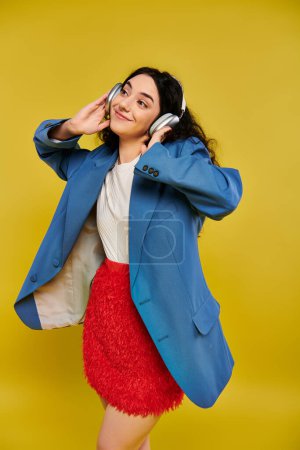 A young, brunette woman with curly hair exudes style and confidence in a blue jacket and red skirt against a vibrant yellow backdrop.