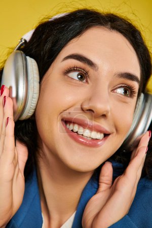 Foto de Una mujer joven y elegante con el pelo rizado sonriendo felizmente a la cámara mientras usa auriculares en un estudio con un fondo amarillo. - Imagen libre de derechos
