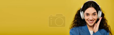 Foto de Mujer joven con el pelo rizado, con un atuendo elegante, sonriendo con auriculares en un estudio con un fondo amarillo. - Imagen libre de derechos