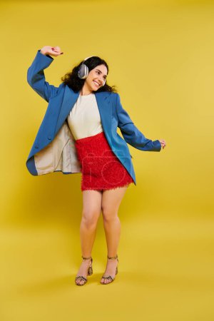 Téléchargez les photos : Une jeune femme brune aux cheveux bouclés émouvant dans une veste bleue élégante et jupe rouge sur un fond jaune vif. - en image libre de droit