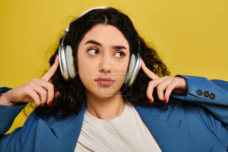 Téléchargez les photos : Une jeune femme élégante aux cheveux bouclés, portant des écouteurs, immergée dans la musique avec une expression sereine sur un fond jaune. - en image libre de droit