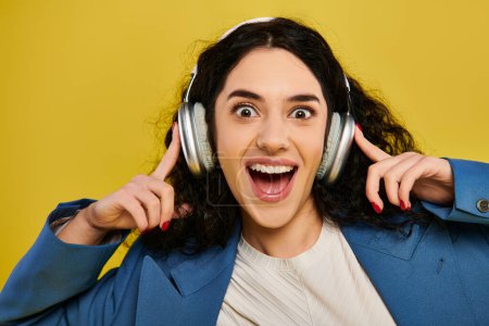 A young, curly-haired brunette woman in stylish attire makes a funny face while wearing headphones in a studio with a yellow background.