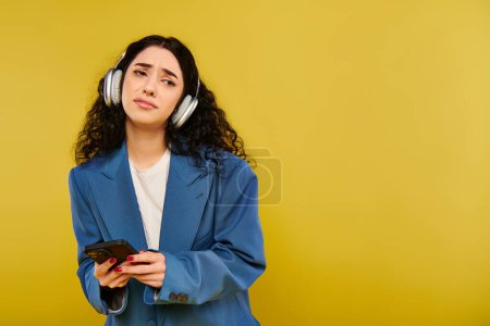 Photo for A brunette woman with curly hair wearing headphones and holding a cell phone, lost in music against a yellow backdrop. - Royalty Free Image