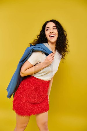 Téléchargez les photos : Une jeune femme aux cheveux bouclés pose dans une jupe rouge vibrante et une chemise blanche, mettant en valeur ses émotions sur fond jaune. - en image libre de droit