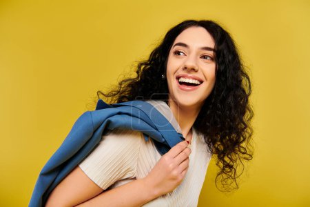 A stylish young woman with curly hair exudes confidence, wearing a striking blue scarf around her neck against a vibrant yellow backdrop.