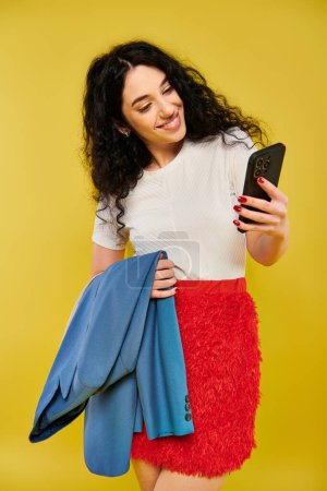 Brunette woman with curly hair in red skirt poses with cell phone in hand, emoting in a studio setting with yellow backdrop.
