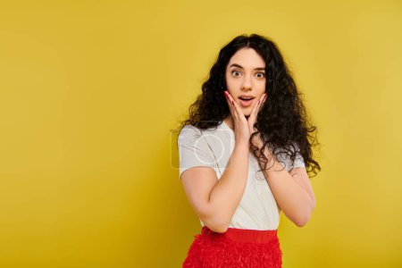 Photo for Young brunette woman with curly hair, wearing a red skirt, expresses emotions by covering her face with her hands. - Royalty Free Image