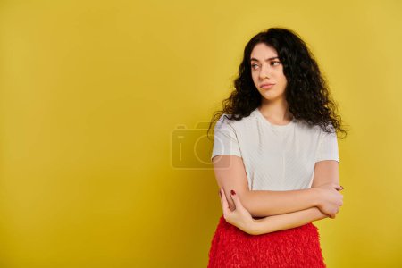 Foto de Una mujer joven y elegante con el pelo rizado se levanta con confianza con los brazos cruzados contra un vibrante telón de fondo amarillo. - Imagen libre de derechos