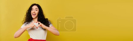 Foto de Young, curly-haired brunette woman in chic attire, forming heart shape with her hands against vibrant yellow backdrop. - Imagen libre de derechos