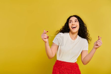 Téléchargez les photos : Une jeune femme brune aux cheveux bouclés pose dans une élégante jupe rouge et chemise blanche, mettant en valeur ses émotions sur un fond jaune. - en image libre de droit