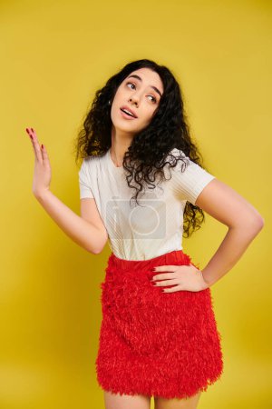 Téléchargez les photos : Une jeune femme brune aux cheveux bouclés pose dans une jupe rouge saisissante, mettant en valeur ses émotions dans un studio au fond jaune. - en image libre de droit