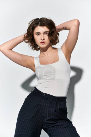 Photo for A young woman in black pants and a white tank top strikes a confident pose, hands elegantly placed on her head. - Royalty Free Image