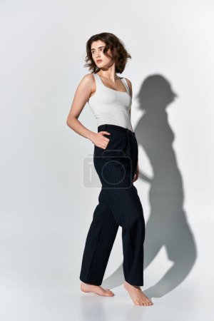 Foto de Una hermosa joven posa con confianza en pantalones negros y una camiseta blanca, de pie con gracia frente a una pared blanca. - Imagen libre de derechos