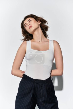 Photo for A pretty young woman striking a pose in a white tank top and black pants, set against a neutral grey backdrop. - Royalty Free Image