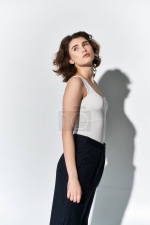A pretty young woman in black pants and white tank top poses elegantly in front of a white wall in a studio setting.