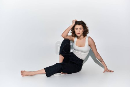 Eine hübsche junge Frau sitzt anmutig mit verschränkten Beinen auf dem Boden, bekleidet mit einer schwarzen Hose und einem weißen Tank-Top.